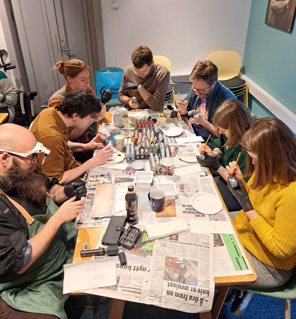 A group of people of different genders gathered around a table painting miniatures.
